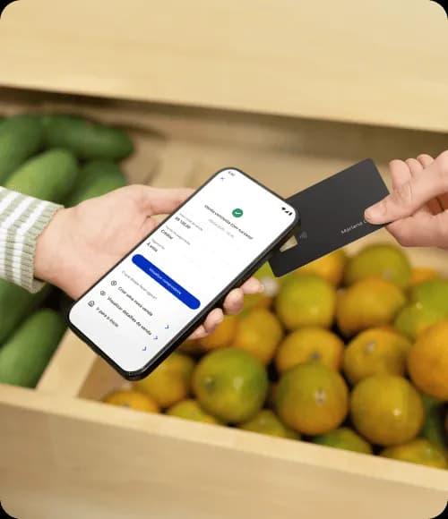 Vendedora de loja de frutas e verduras segurando um celular enquanto um cliente aproxima seu cartão de crédito